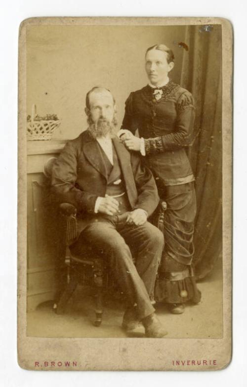 Seated bearded man with lady standing beside him.