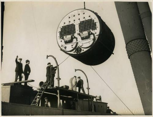 Photograph of eight men onboard a ship with a Scotch boiler being hoisted