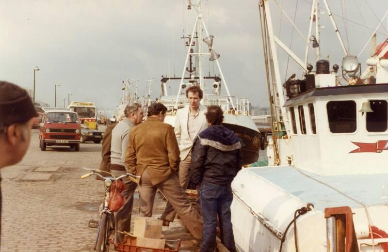 Colour Photograph Showing Group Of Fishermen Chatting On Quayside, One With Bicycle