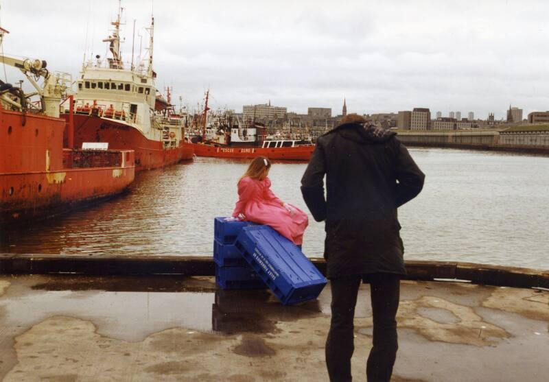 Colour Photograph Showing A Small Girl On Empty Fish Boxes At The Harbour, With Photographer