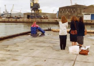 Colour Photograph Showing Small Girl On Empty Fish Boxes, With Women Looking On, At Harbour